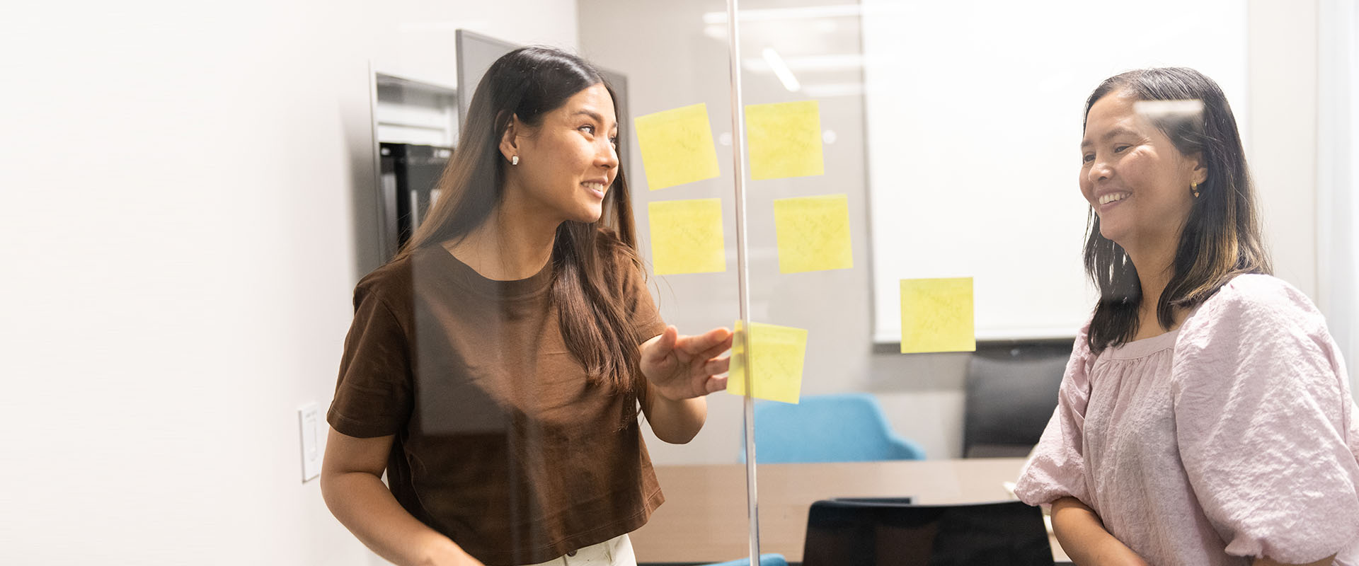 Two professionals smile while collaborating in a modern office, placing yellow sticky notes on a glass partition to brainstorm project tasks.