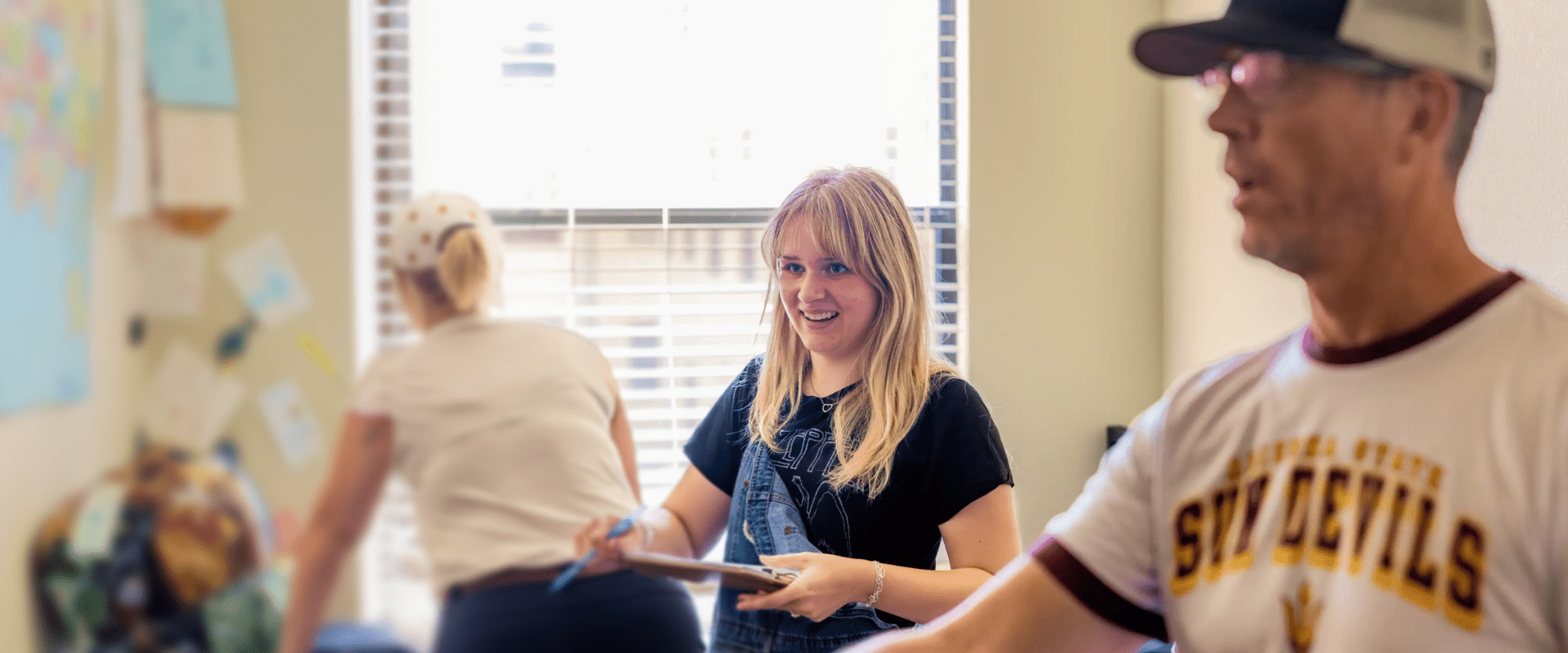 A young woman with blonde hair, wearing overalls and a black t-shirt, smiles while holding a clipboard in a classroom or office. In the foreground, a man wearing an Arizona State Sun Devils t-shirt is partially visible, while another woman works in the blurred background near a window.
