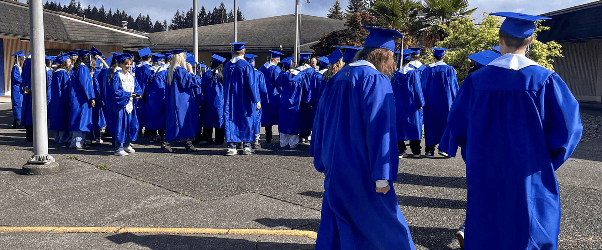 A large group of graduating students wearing bright blue graduation gowns and matching mortarboard caps stands outside on a paved courtyard near school buildings. Most students have their backs turned or are chatting in small groups under a bright, sunny sky.