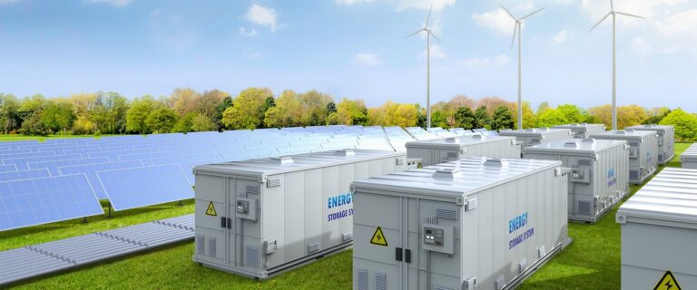 Large white modular energy storage battery containers lined up in a field alongside solar panels and wind turbines under a blue sky.