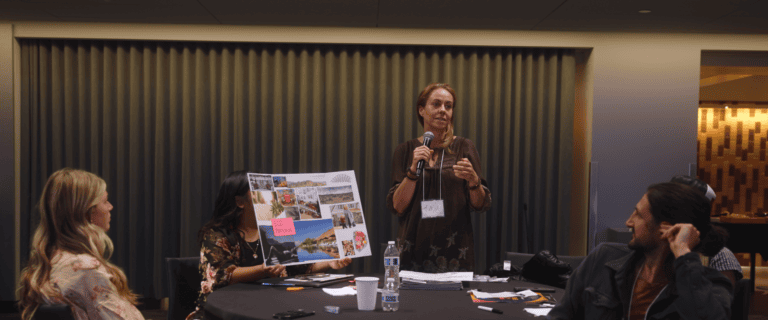 A woman wearing a brown top and a name tag that reads "Karla" speaks into a handheld microphone while facilitating a workshop. Seated participants surround a round table, with one person holding up a collage poster board featuring landscape and lifestyle photos alongside a pink sticky note labeled "ECO PERSONA."
