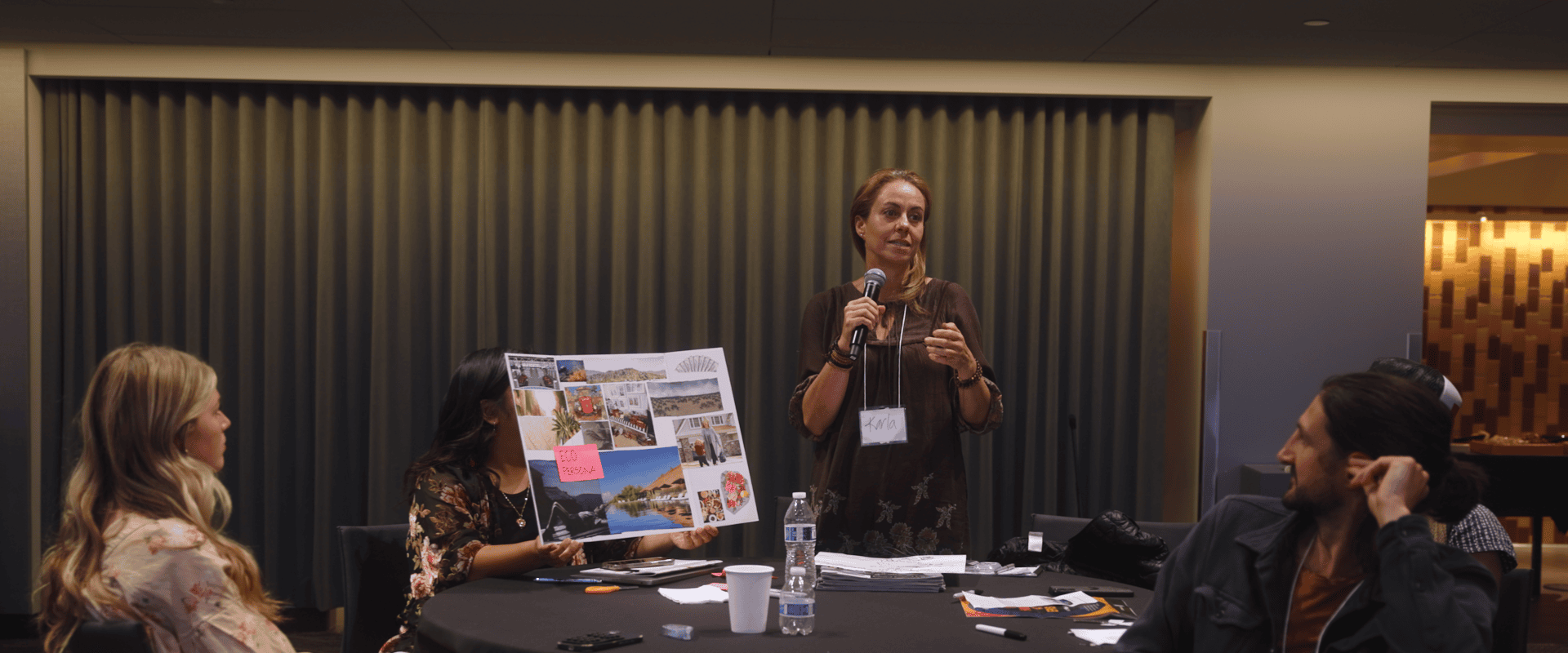 A woman wearing a brown top and a name tag that reads "Karla" speaks into a handheld microphone while facilitating a workshop. Seated participants surround a round table, with one person holding up a collage poster board featuring landscape and lifestyle photos alongside a pink sticky note labeled "ECO PERSONA."