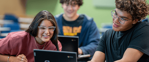 A young person with long dark hair, clear-framed glasses, and a pink shirt leans over to look at a laptop screen alongside another young person with curly hair and glasses in a black t-shirt. A third person sits blurred in the background working on a laptop.