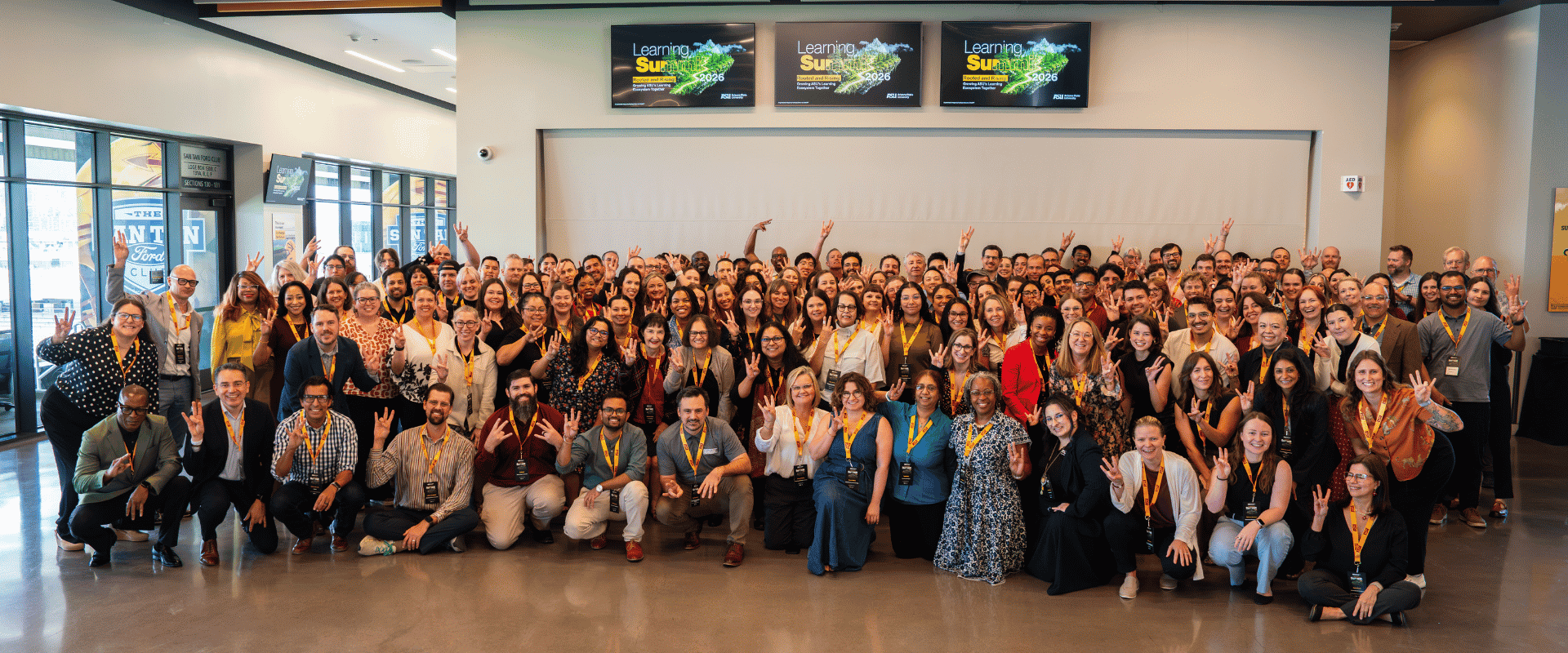 A large, diverse group of attendees gathers indoors for a group photo at the ASU Learning Summit 2026. Everyone is smiling, with many people making the Arizona State University "Fork 'Em" hand gesture. Three digital display screens hanging overhead show the "Learning Summit 2026" event branding.